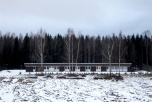 En låg, brun och vit byggnad mellan en snötäckt åker och en mörk skog